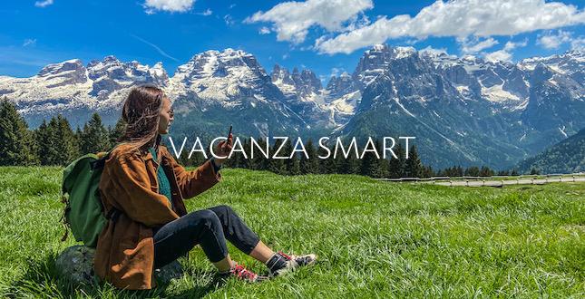 A person sitting on grass, taking a photo of snow-capped mountains under a blue sky, with "VACANZA SMART" text.