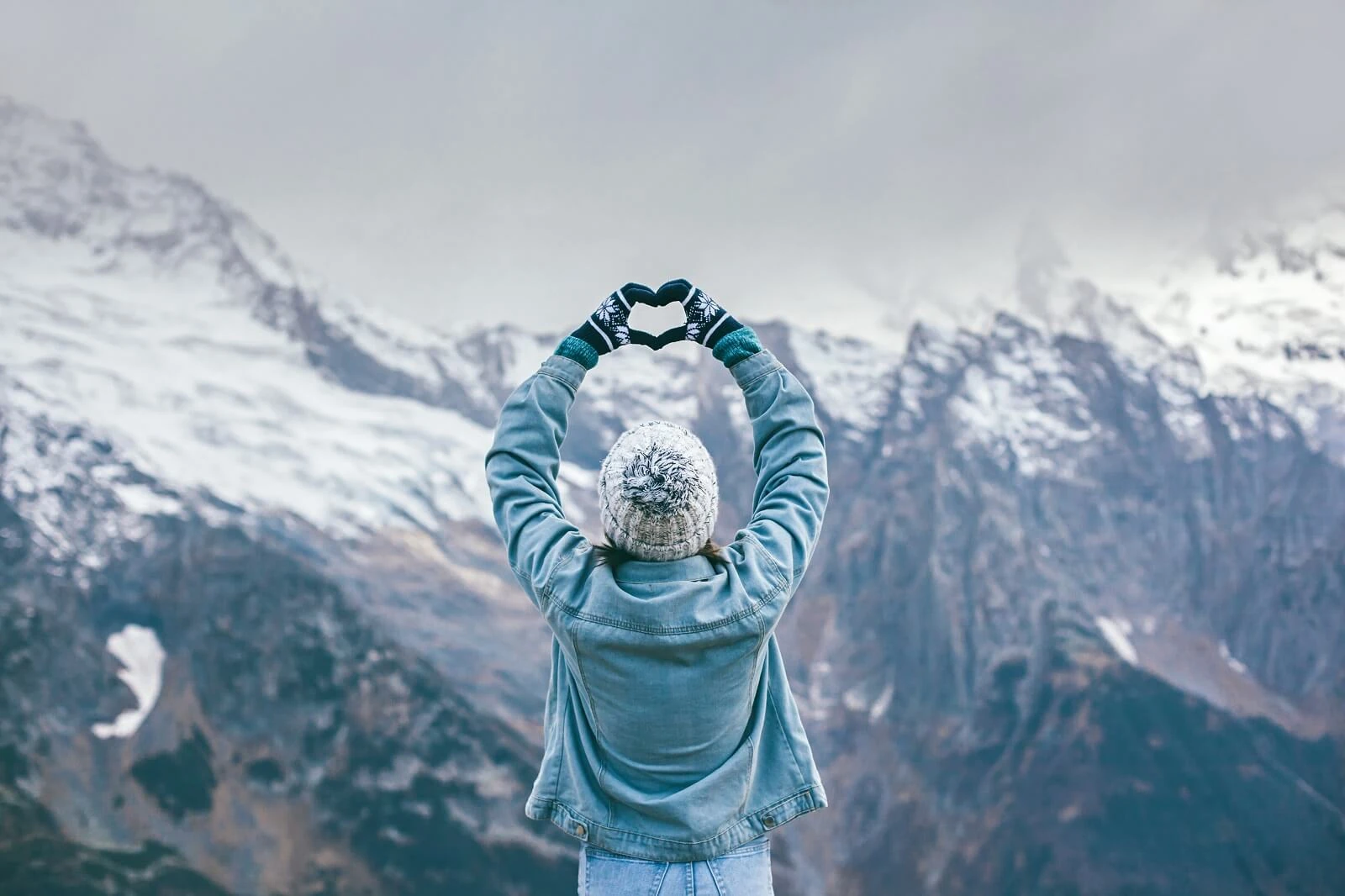 Person in a winter outfit forming a heart shape with hands against a snowy mountain backdrop.