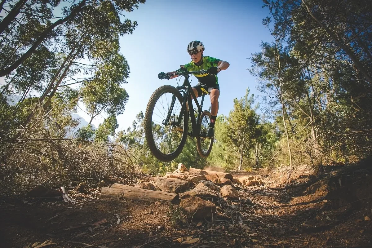 A cyclist performs a jump on a mountain bike trail surrounded by trees.